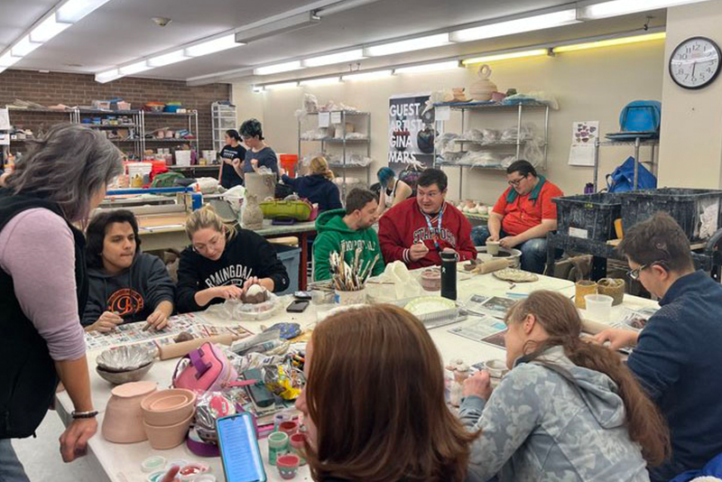 Un profesor observa los trabajos que varios alumnos de arte están realizando en una mesa de una gran sala.