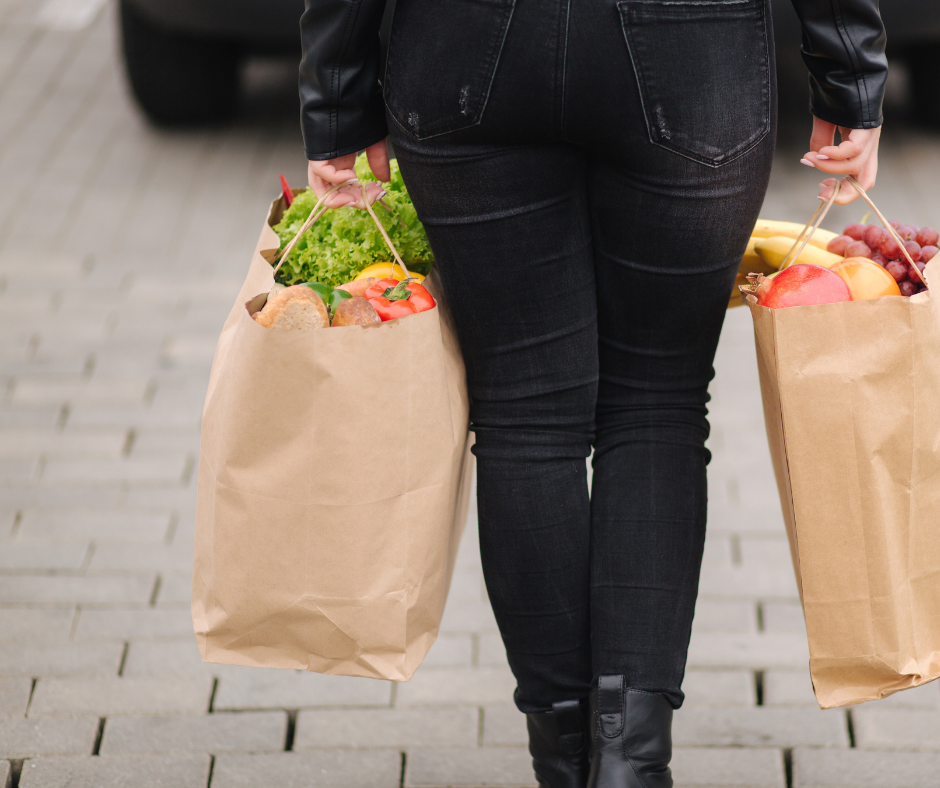 A view of a woman in black jeans from behind carrying a grocery bag in each hand.