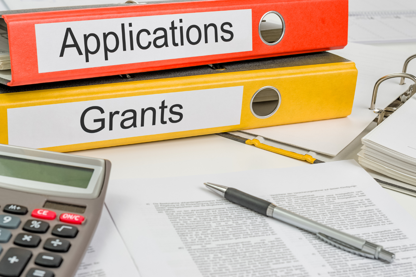 two binders appear on a desk with papers, pen and a calculator. One says "Applications" the other says "Grants."