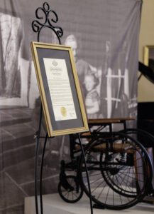 A golden framed document sits on an easel in front of an antique wheelchair.