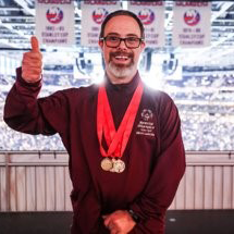 Matt Schuster a young white man with a beard and glasses appears with his thumb up wearing three gold medals.