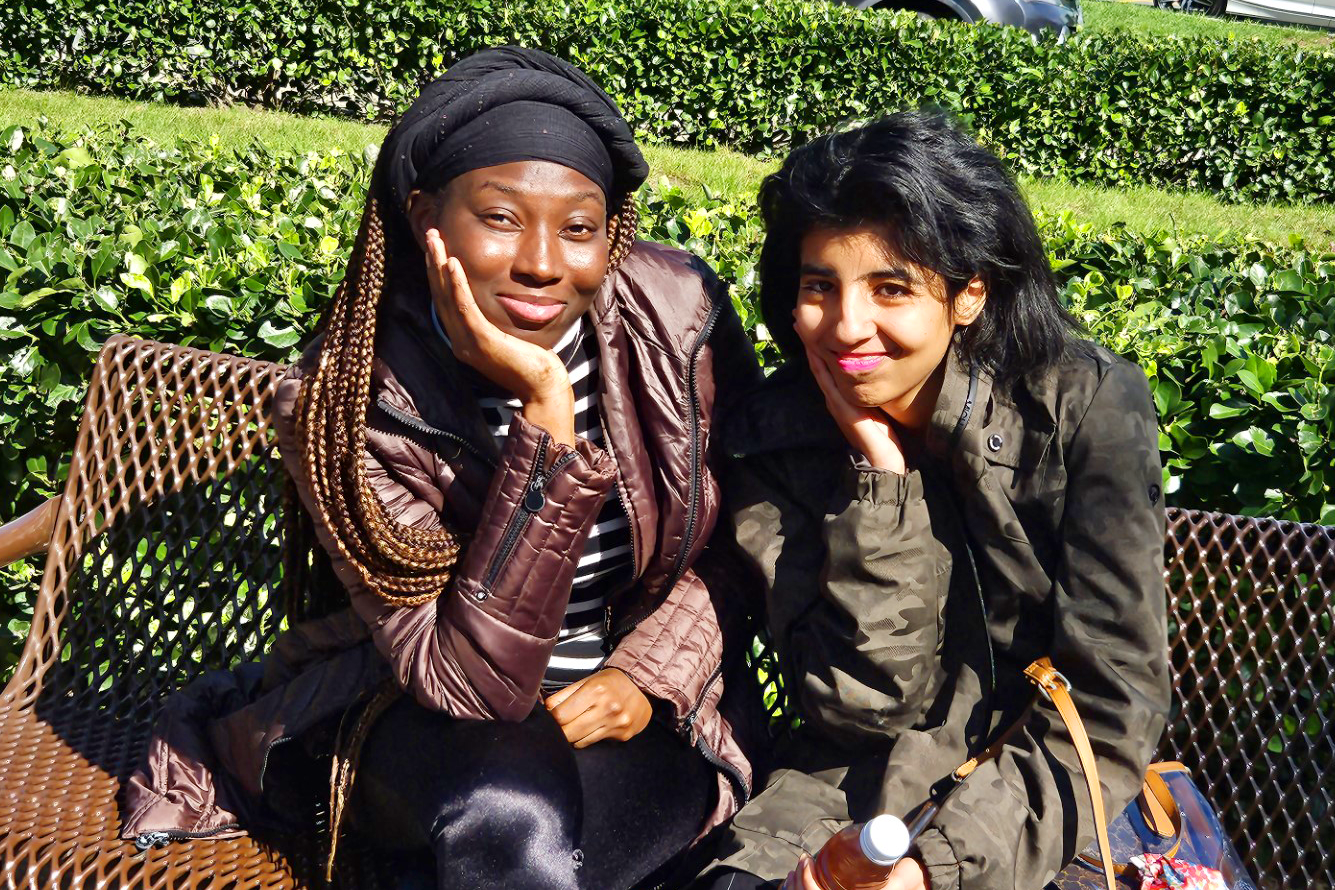 Two young independent women: A young black woman sits next two a young brown woman on a bench in a park. Both have the heads resting on their hands in the same fashion.