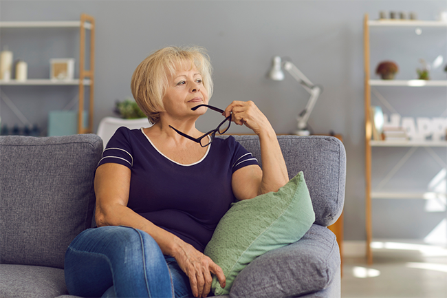 Self-Direction: A middle aged woman is considering her decision-making options on a couch holding her glasses.