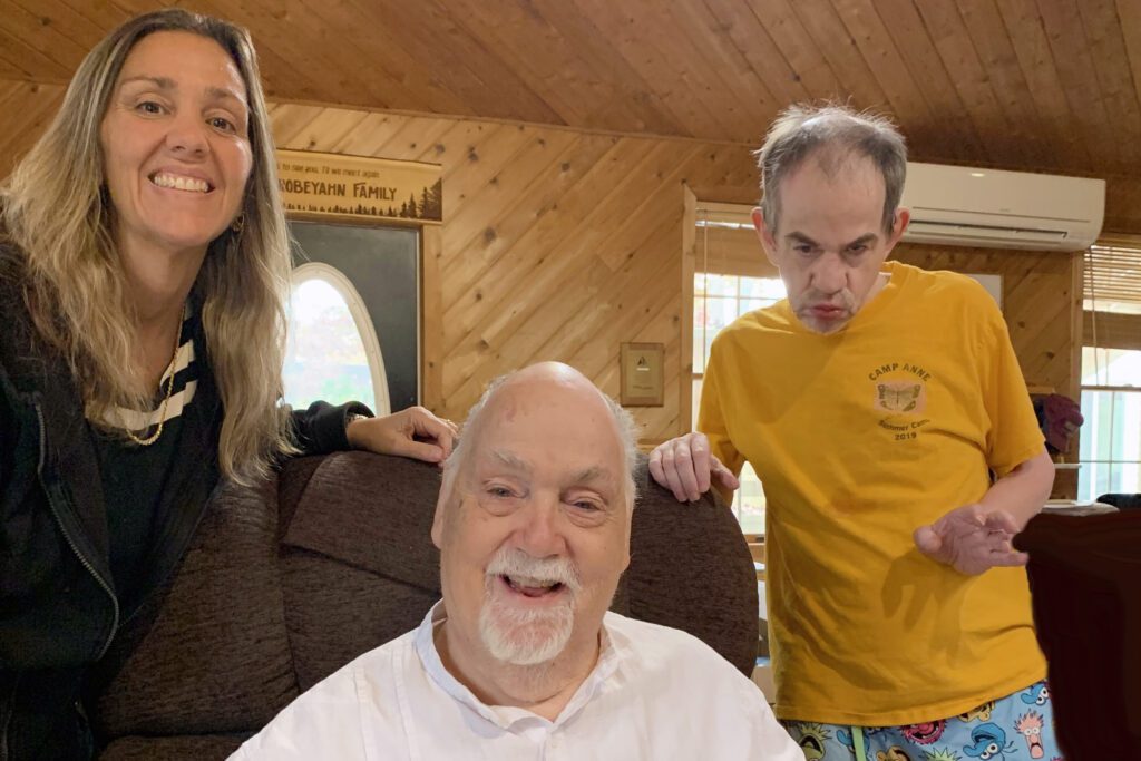 Care manager is a partner: Care Manager is a woman to the left smiling at the camera with an elderly man in a chair center and to the right is a middle-aged man with autism.