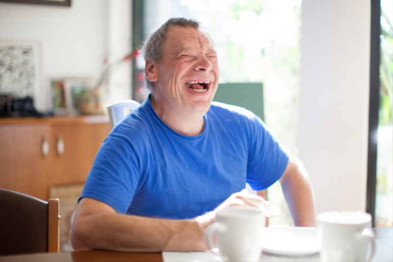 An older man with Down syndrome is laughing at a table in the sunshine.