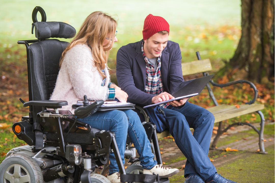 A young woman in a wheelchair looks at a tablet with a young man who is sitting on a park bench.