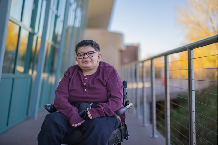 A young person with a purple shirt smiles, sitting in a wheelchair.